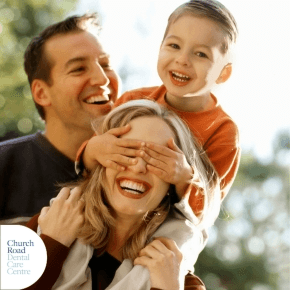 A man and a woman smiling and a young boy on the woman's shoulders with his hands over her eyes