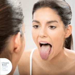 A lady with dark hair in a ponytail looking into a mirror sticking her tongue out fully